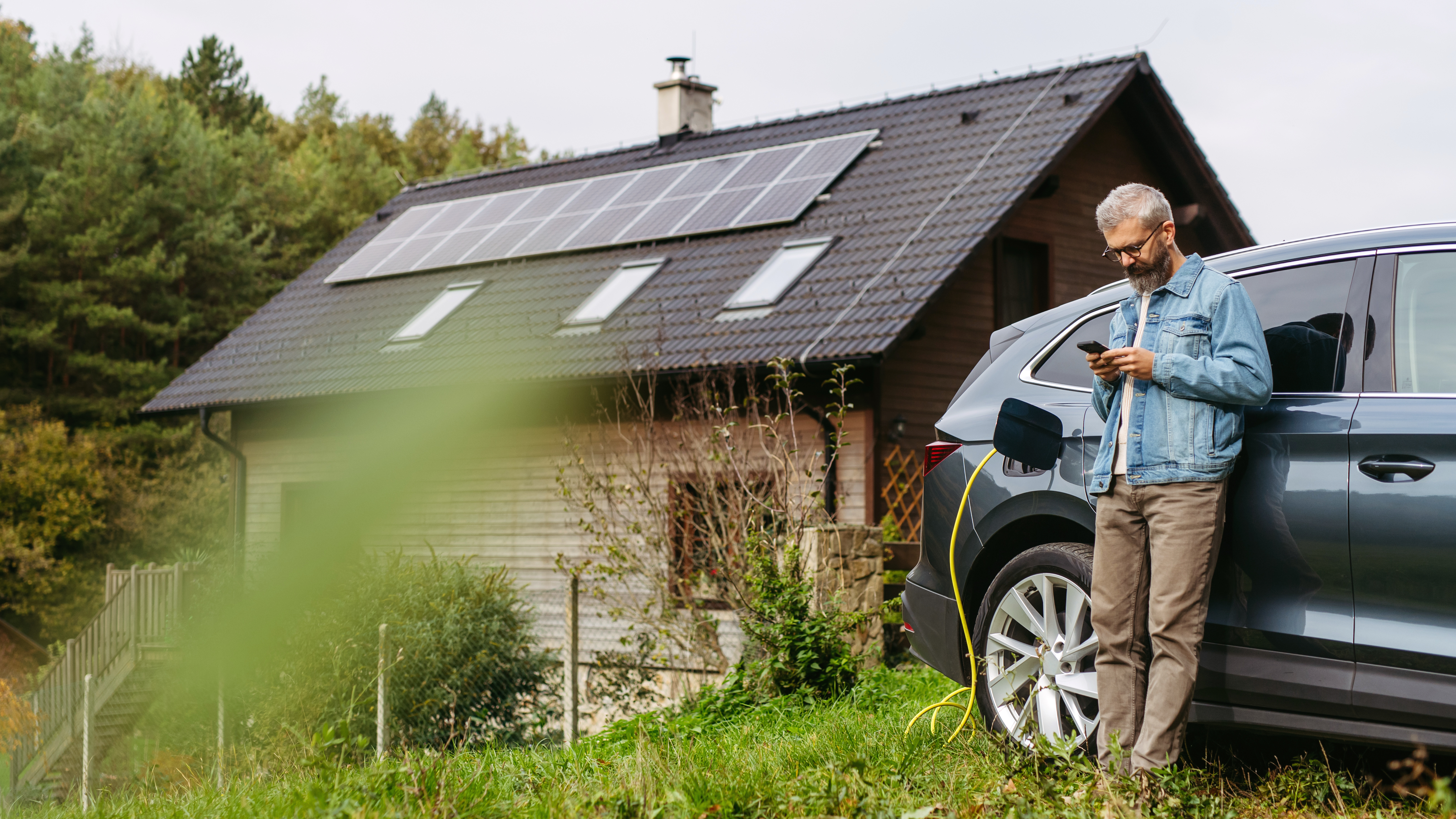 Mann lädt Elektroauto vor seinem Haus mit Solaranlage auf dem Dach hinter sich. Mann, der sich an ein Auto lehnt, während er auf dem Smartphone scrollt.